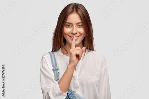 Shh, its privacy! Good looking dark haired woman with charming smile, keeps index finger over mouth, shows hush gesture, gossips with best friend about boyfriends, dressed in fashionable clothes
