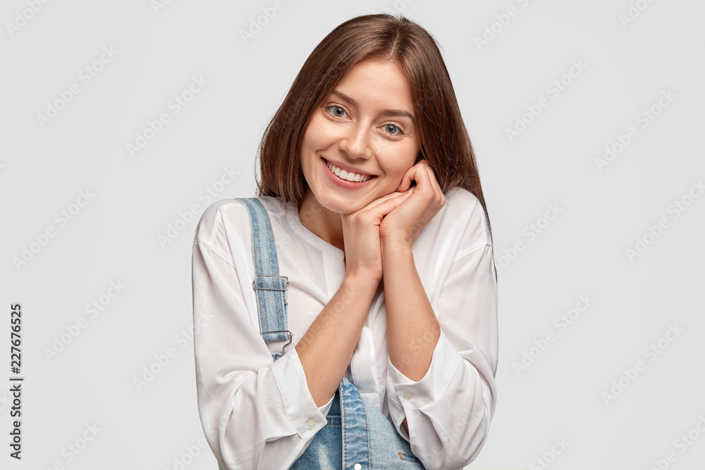 Content young female keeps hands near cheek, smiles positively, expresses happiness and truthful feelings, dressed in casual outfit, poses against white background, says: Mmm, its so pleasant! Content young female keeps hands near cheek, smiles positively, expresses happiness and truthful feelings, dressed in casual outfit, poses against white background, says: Mmm, its so pleasant!