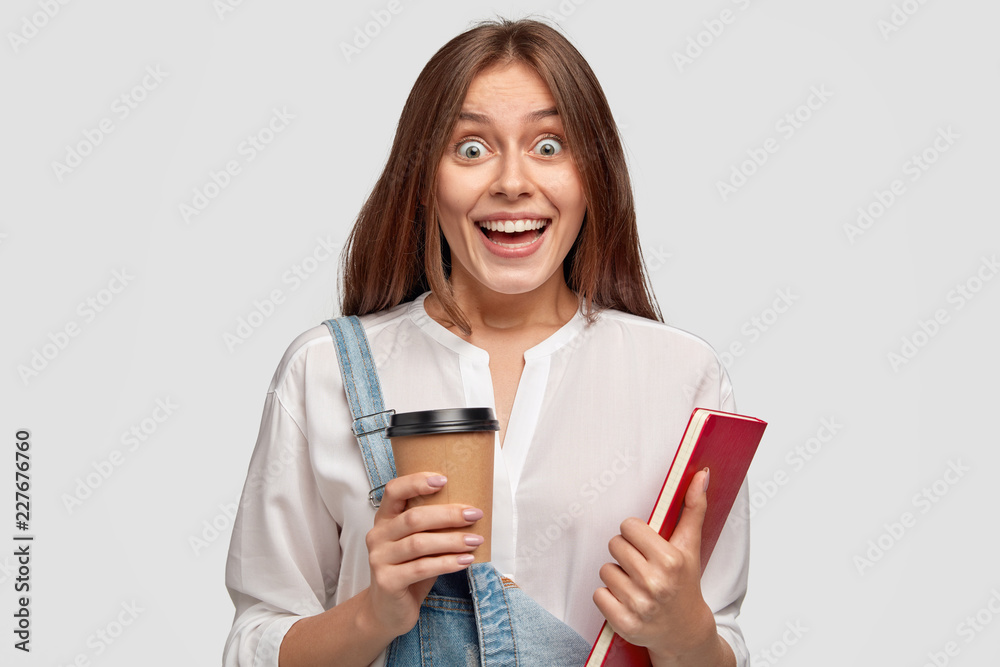 Emotional cheerful student has surprised expression, smiles broadly, drinks takeaway coffee and notepad, hears something unexpected, isolated over white background. Happy schoolgirl models indoor Emotional cheerful student has surprised expression, smiles broadly, drinks takeaway coffee and notepad, hears something unexpected, isolated over white background. Happy schoolgirl models indoor