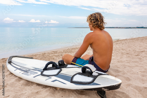 Handsome man sits on the beach with white blank surfing board wait for wave to surf spot at sea ocean shore. Concept of sport, fitness, freedom, happiness, new modern life, hipster. © Georgii