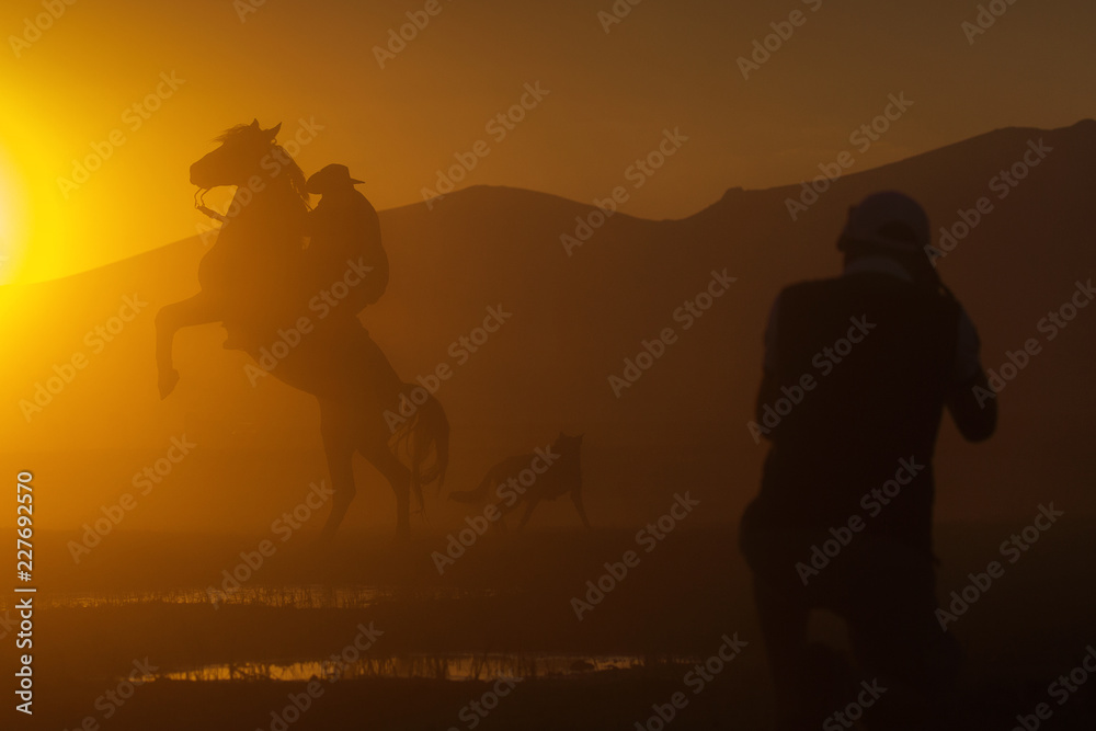 Obraz premium Silhouette of a cowboy puting his horse to stay in two feets at sunset with dust in background and a photographer taking pictures of him