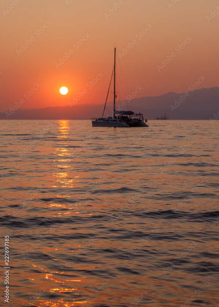 Yacht in the sea at sunset. Beautiful orange sunset colors, mountains in the background. Calm and tranquility. Holidays in Greece.
