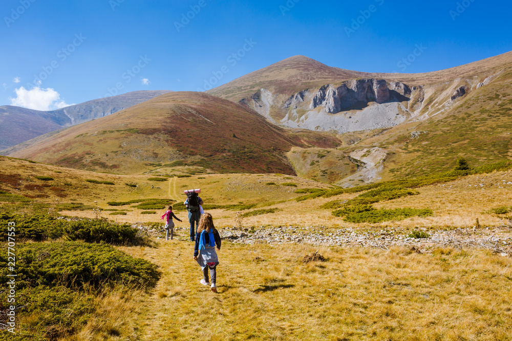 Two little girls and a man walking on a mountain meadow in autumn. Mountain climbing, with backpacks, Grass is yellow, hay. Family hiking up hill, towards the mountain peak. Shot from the back, rear.