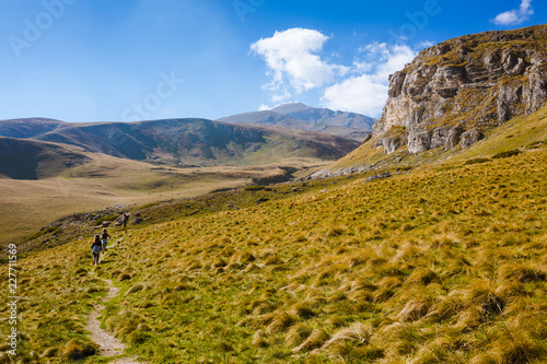 Two little girls and a man walking on a mountain meadow in autumn. Mountain climbing, with backpacks, Grass is yellow, hay. Family hiking up hill, towards the mountain peak. Shot from the back, rear.