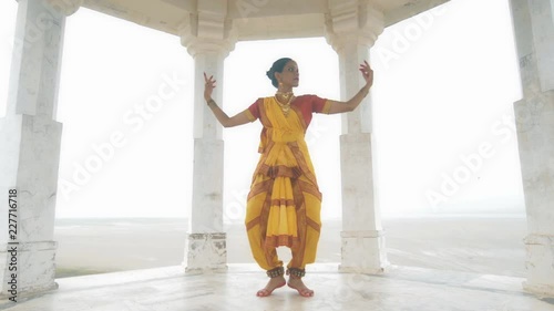 A wide shot of a Indian folk dancer dancing elegantly in a white marble temple located on a hilltop. A movement shot of a beautiful woman in traditional yellow sari performing classical dance form photo