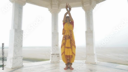 A movement shot of a beautiful woman in traditional yellow sari performing classical dance form. A wide shot of a Indian folk dancer dancing elegantly in a white marble temple located on a hilltop. photo