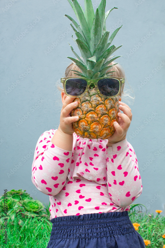 Little girl holding a pineapple with glasses in front of her face. Pineapple  instead of the head. In the garden outdoors. Stock Photo | Adobe Stock