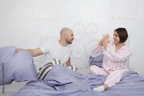 Young beauty and a bald bearded man in pajamas are cursing and quarreling in bed with pillows and blue bedding.