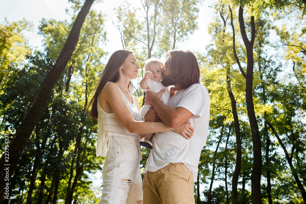 Happy family on the open air. Young dark-haired woman and her husband are kissing their charming little daughter. Happy family on the open air. Young dark-haired woman and her husband are kissing their charming little daughter.
