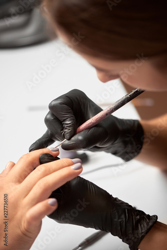Process of making fashionable manicure in beauty salon. Close-up profile of professional master manicurist applying pastel varnish on client woman nails using brush on light blurred background.