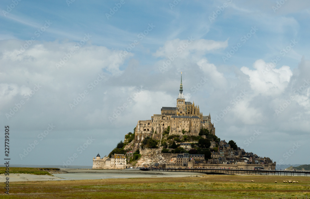 Beautiful view of famous historic Le Mont Saint-Michel abbey tidal island on fields of fresh green grass on a sunny day with blue sky and clouds in summer, Normandy, northern France