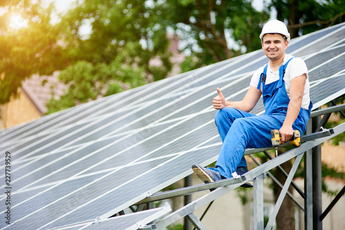 Young smiling electrician sitting on almost finished stand-alone solar photo voltaic panel system with screwdriver, showing thumb up gesture on bright sunny green tree background © anatoliy_gleb
