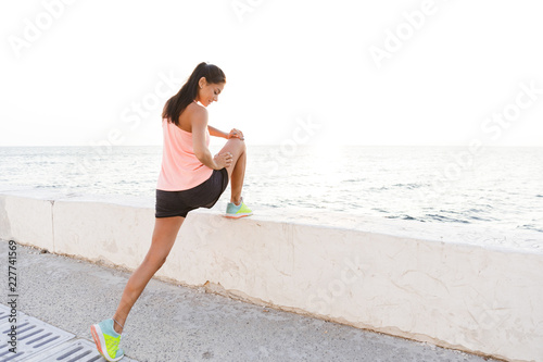 Photo of runner woman 20s in tracksuit stretching legs, during doing sports at seaside