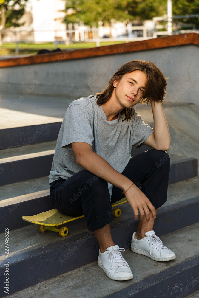 Photo of caucasian skater boy 16-18 in casual wear sitting on skateboard in  skate park, during sunny summer day Photos | Adobe Stock