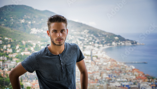 Handsome Young Man in Trendy Attire, in a Sunny Summer Day with Italian Sea Coast in the Distance, Wearing a White Shirt