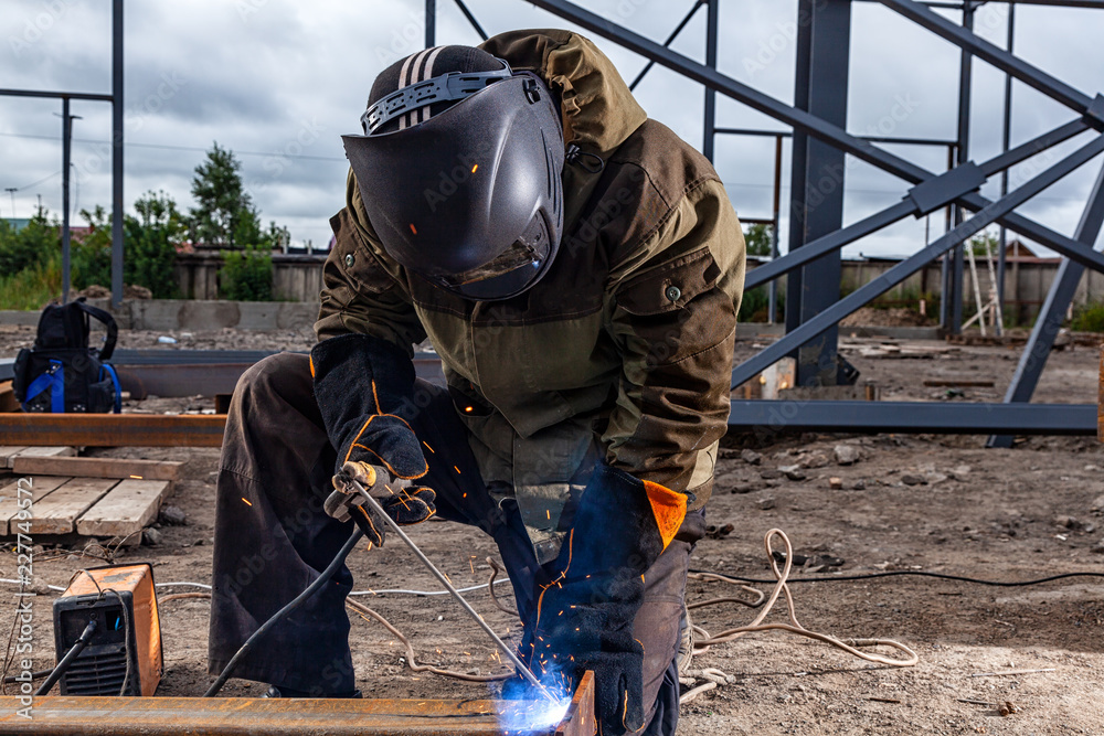 Close up welder weld metal pipe at construction site. A builder in brown uniform, welding mask and welders leathers, weld metal with a arc welding machine, blue sparks fly to the sides Close up welder weld metal pipe at construction site. A builder in brown uniform, welding mask and welders leathers, weld metal with a arc welding machine, blue sparks fly to the sides