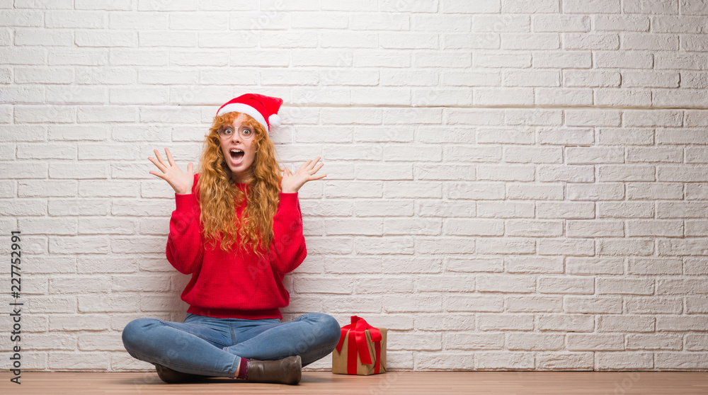 Young redhead woman sitting over brick wall wearing christmas hat celebrating crazy and amazed for success with arms raised and open eyes screaming excited. Winner concept Young redhead woman sitting over brick wall wearing christmas hat celebrating crazy and amazed for success with arms raised and open eyes screaming excited. Winner concept