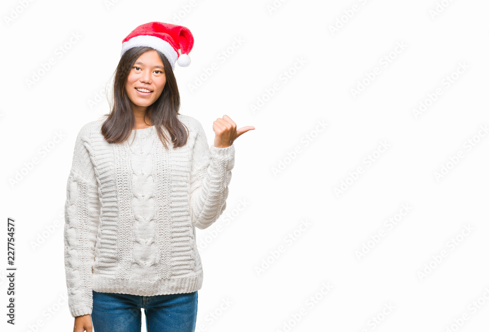 Young asian woman wearing christmas hat over isolated background smiling with happy face looking and pointing to the side with thumb up. Young asian woman wearing christmas hat over isolated background smiling with happy face looking and pointing to the side with thumb up.