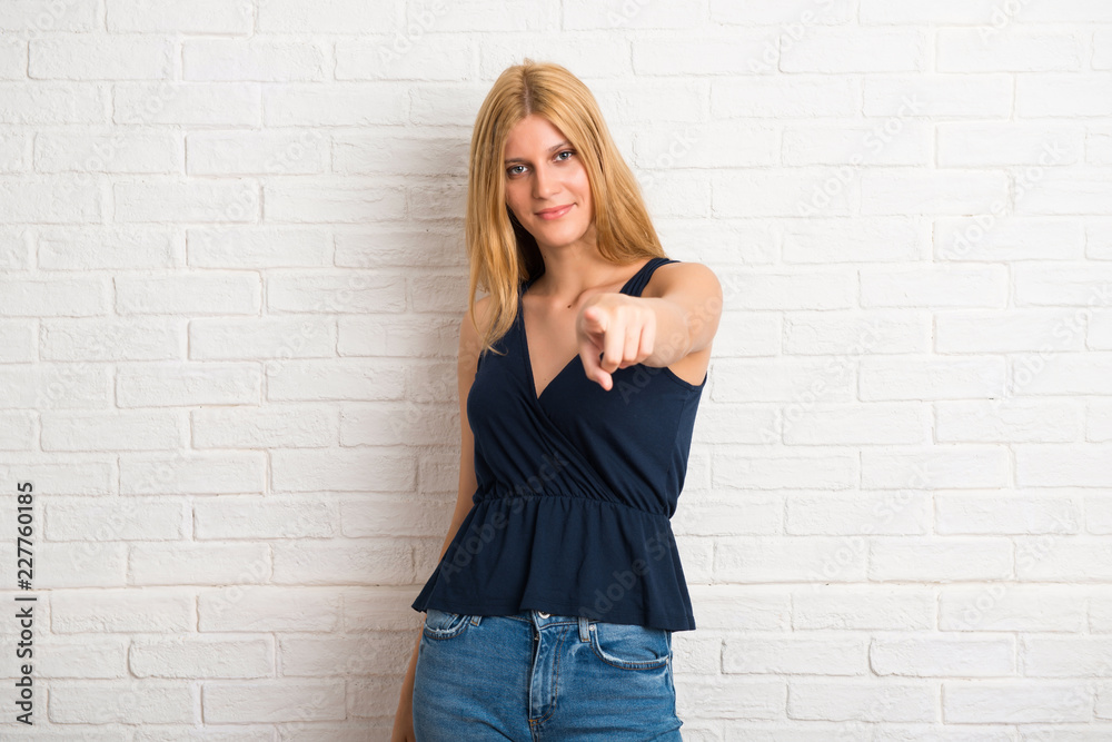 Blonde woman points finger at you with a confident expression on white brick wall background Blonde woman points finger at you with a confident expression on white brick wall background