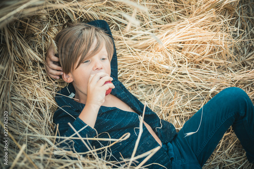 Fair-haired boy lies on hay background and eats an apple. Sale for entire autumn collection, incredible discounts and wonderful choice. Back to school. Bye summer - hi autumn.