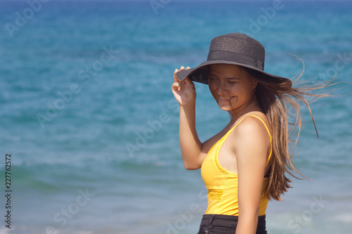 Young dark-haired beautiful European girl in black hat and yellow swimsuit smiling and standing on the background of the sea and clouds on a Sunny day