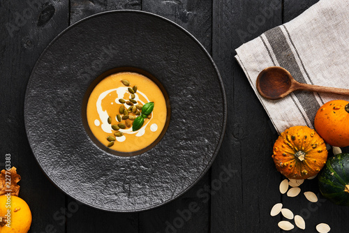 Pumpkin and carrot soup with pumpkin seeds, cream and basil in black modern plate on dark wooden background. Top view.