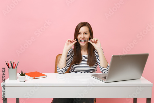 Passionate woman in casual clothes gnawing hold pencil in teeth sit work at white desk with contemporary pc laptop isolated on pastel pink background. Achievement business career concept. Copy space.