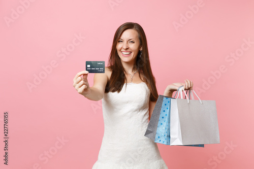 Cheerful bride woman in white wedding dress holding credit card and multi colored packages bags with purchases after shopping isolated on pink background. Organization of wedding concept. Copy space.