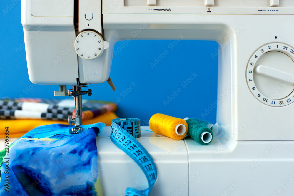 Sew clothes on a sewing machine. Sewing clothes made of blue silk on a blue  background. Sewing machine, measuring tape, thread and lightweight fabric  for sewing clothes. Stock Photo