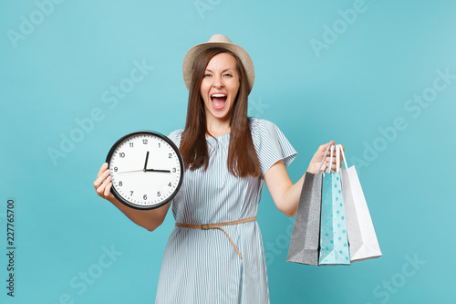 Portrait beautiful young caucasian woman in summer dress, straw hat holding packages bags with purchases after shopping, round clock isolated on blue pastel background. Copy space for advertisement.