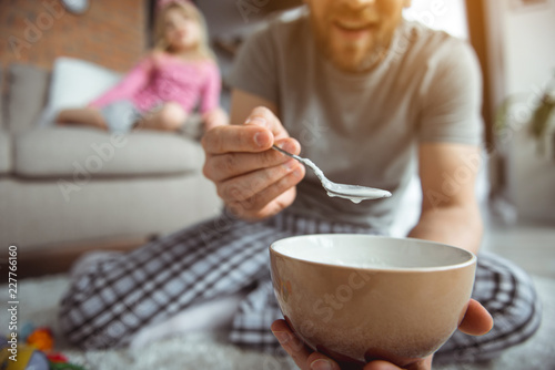 Eat one more spoon.  Close up of caring dad is feeding his baby by porridge. She is holding bowl and smiling. Girl is sitting on sofa on background. Focus on spoon 