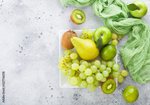 Fresh raw organic green toned fruit on white ceramic square plate on stone kitchen table background. Pear and grapes with kiwi and lime and apples. Space for text. Top view