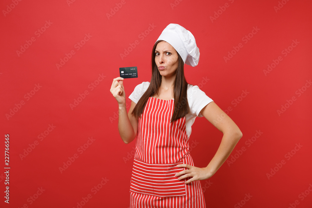 Housewife female chef cook baker in striped apron white t-shirt toque chefs hat isolated on red wall background. Confused woman hold in hand credit bank card cashless money. Mock up copy space concept Housewife female chef cook baker in striped apron white t-shirt toque chefs hat isolated on red wall background. Confused woman hold in hand credit bank card cashless money. Mock up copy space concept