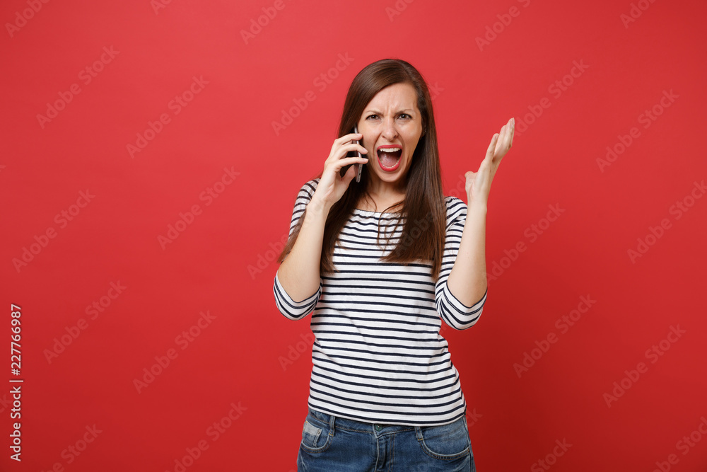Angry young woman screaming, spreading hands, talking on mobile phone, conducting conversation isolated on bright red wall background. People sincere emotions, lifestyle concept. Mock up copy space. Angry young woman screaming, spreading hands, talking on mobile phone, conducting conversation isolated on bright red wall background. People sincere emotions, lifestyle concept. Mock up copy space.