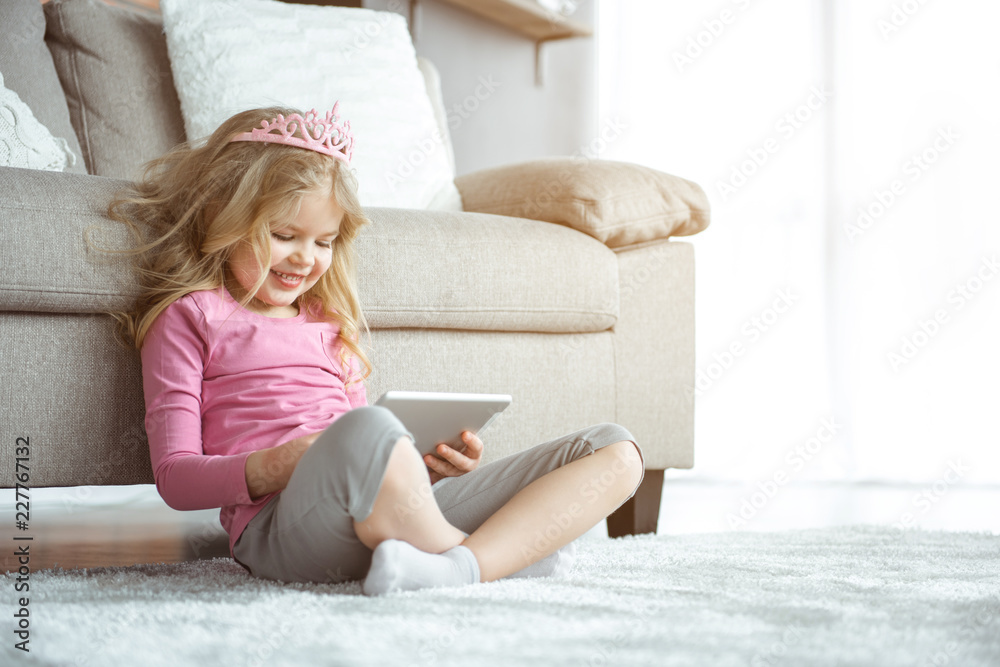 Adorable little girl is entertaining with gadget at home. She is touching screen and laughing. Child is sitting on floor near couch and wearing pink crown. Copy space Adorable little girl is entertaining with gadget at home. She is touching screen and laughing. Child is sitting on floor near couch and wearing pink crown. Copy space