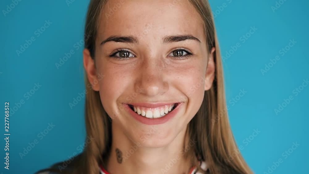Close up of a blonde cute girl face. Girl is looking in the camera and smiling. Portrait shot. Attractive young teenage girl smiling at camera in indoors on the blue wall background