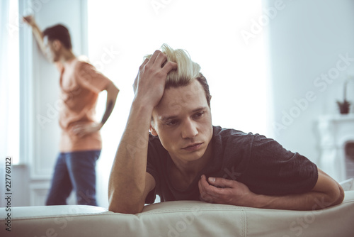 Feeling offended. Toned portrait of sad guy with dyed hair sitting on couch while his boyfriend standing near window on blurred background