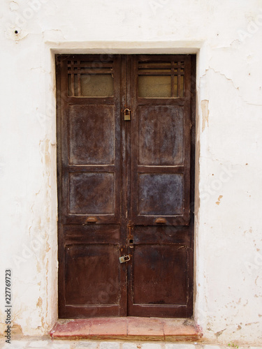 old wooden brown double doors with chipped flaking faded peeling paint and rusty handles padlocked closed in a white painted wall and tile doorstep
