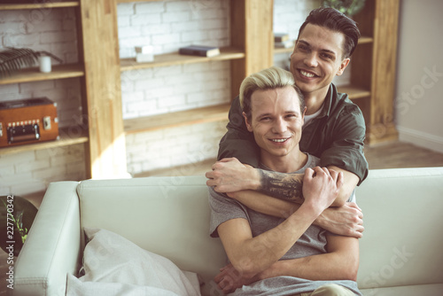 Toned waist up portrait of loving gay couple cuddling at home. They looking at camera with happy smiles