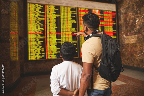 Rear view of hindu travelers standing at the railway station while having a look at the arrival departure board