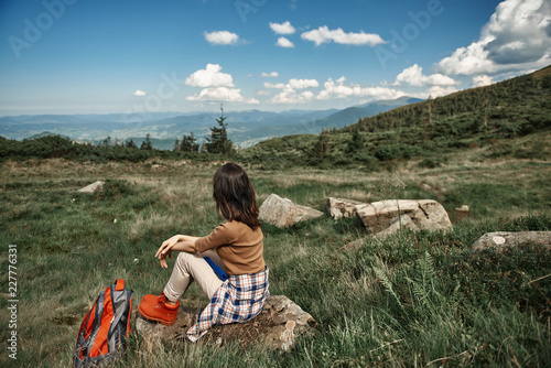 Woman sitting on rock in highland valley during her trip with rucksack. She looking at beautiful view in distance. Copy space in right side
