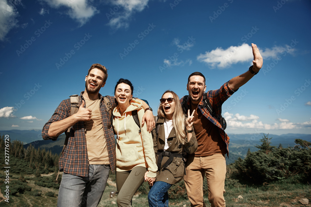 Low angle waist up portrait of jolly two men and two women standing and embracing. Tourists are laughing and raising arms for showing funny gestures