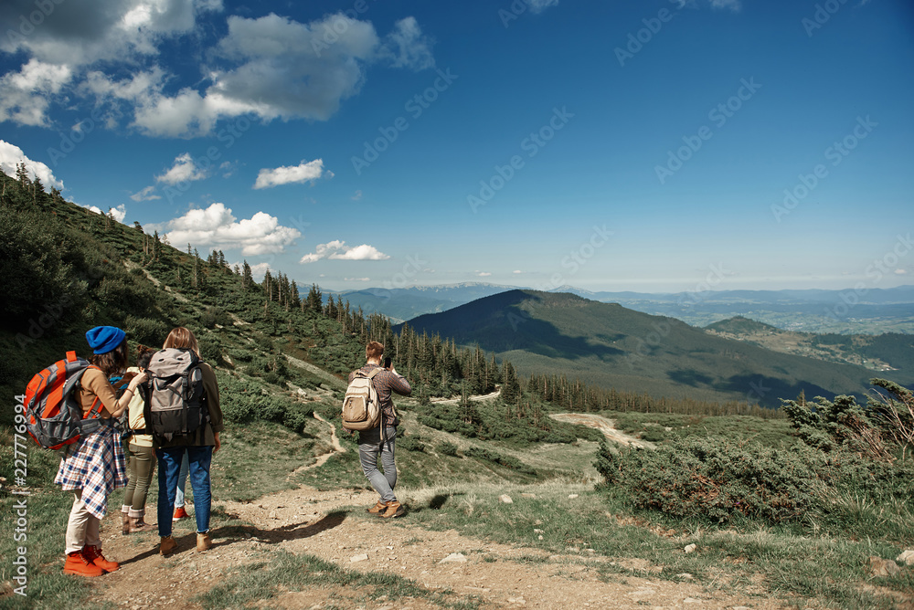 Three women and man travelling among high peaks with rucksacks. They standing and enjoying beautiful view while guy is taking photo of highland with smartphone