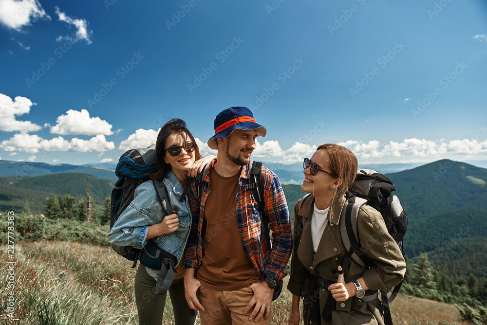 Smiling two women and man backpacking together in mountainous area. They are standing and enjoying beautiful view