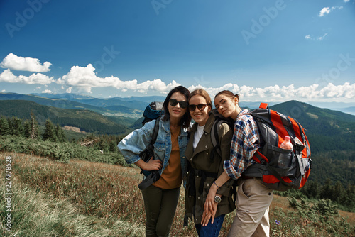 Waist up portrait of three smiling women exploring mountainous landscapes together. They standing jointly and embracing while carrying backpacks with tourist stuff