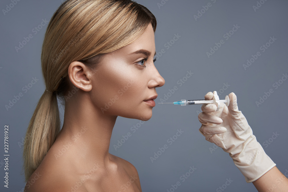Laconic photo on the grey background with young lady sitting and getting injection into her lip Laconic photo on the grey background with young lady sitting and getting injection into her lip
