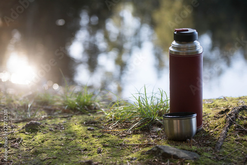 A red thermos and a coffee mug standing in the moss beside a lake in the autumn, outdoor living background