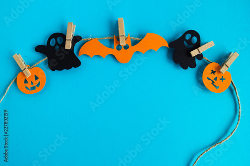 Holiday decorations for Halloween. Black paper ghosts and orange paper pumpkins and bat hanging on a rope on a blue background with copy space.