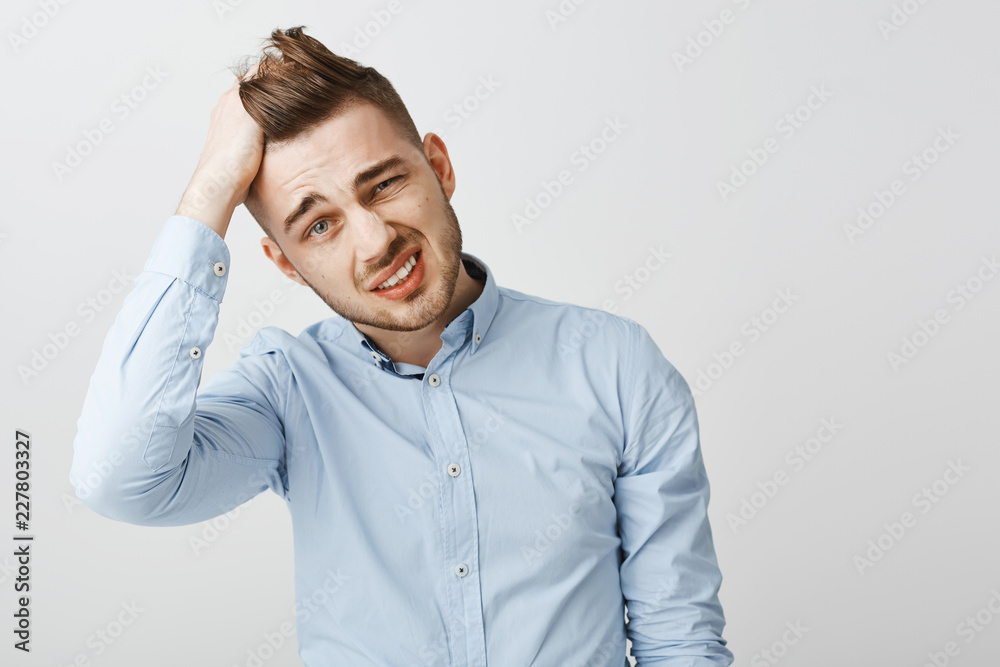 Waist-up shot of perplexed and troubled handsome young businessman in blue stylish shirt tilting head rubbing hair, grimacing and squinting being in terrible tough situation over gray wall Waist-up shot of perplexed and troubled handsome young businessman in blue stylish shirt tilting head rubbing hair, grimacing and squinting being in terrible tough situation over gray wall