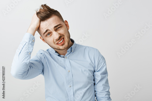 Waist-up shot of perplexed and troubled handsome young businessman in blue stylish shirt tilting head rubbing hair, grimacing and squinting being in terrible tough situation over gray wall